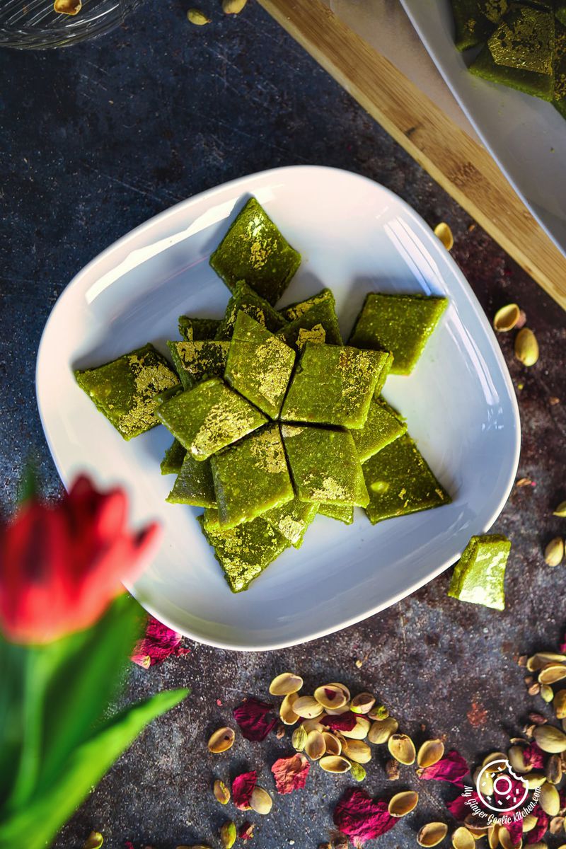 overhead view of golden leaves garnished pista kaju katli in a white plate with some pistachio shells on the side