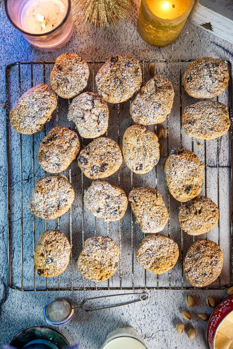 Close-up of pistachio cranberry cookies on a cooling rack with various baking ingredients in the background.