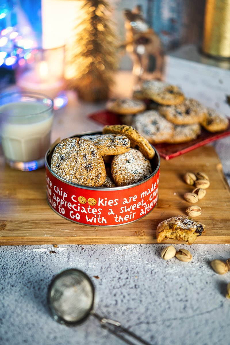 A stack of pistachio cranberry cookies dusted with powdered sugar on a red plate, with milk and holiday lights in the background.