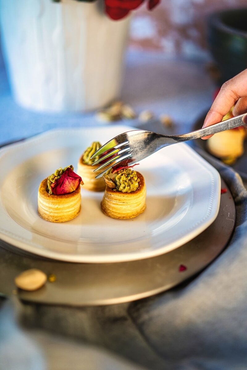 Three no-bake pistachio cream cups on a plate, decorated with edible rose petals, with a fork about to pick one up