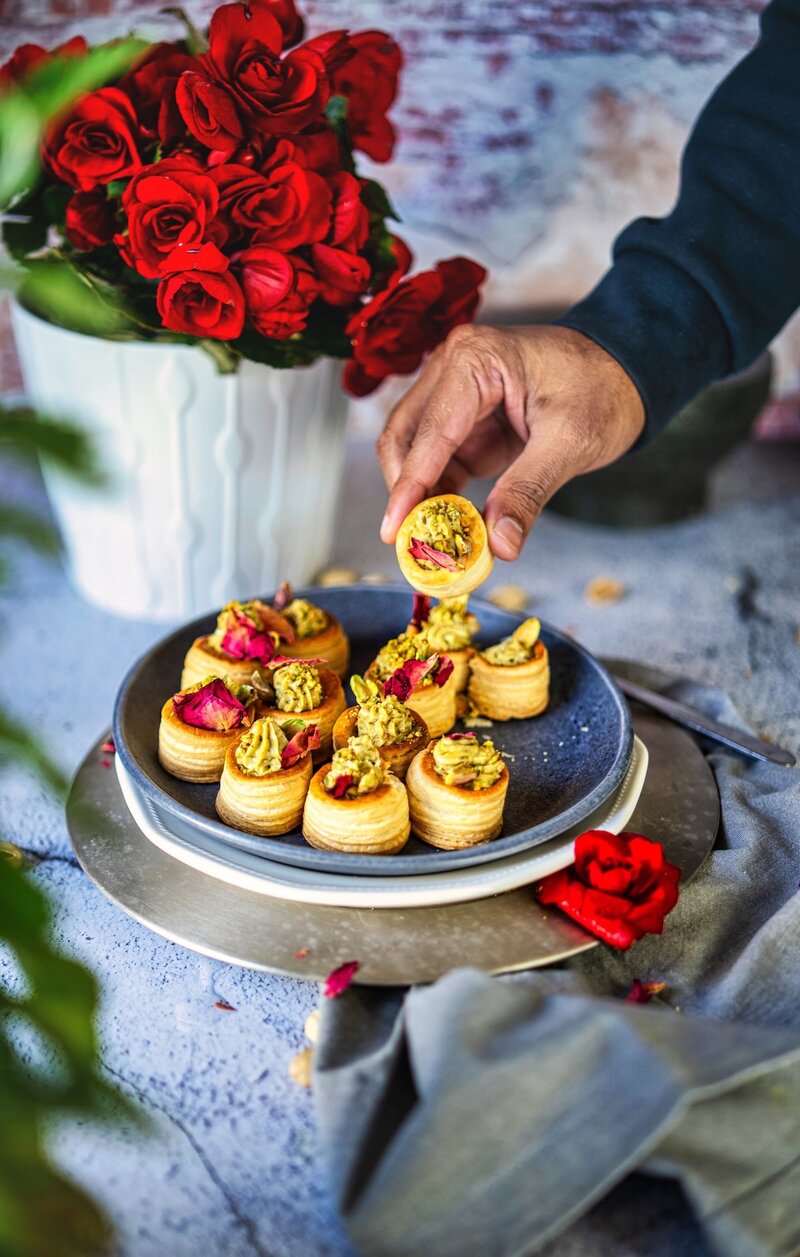 Hand picking up a no-bake pistachio cream cup from a plate, surrounded by rose petals and fresh red roses