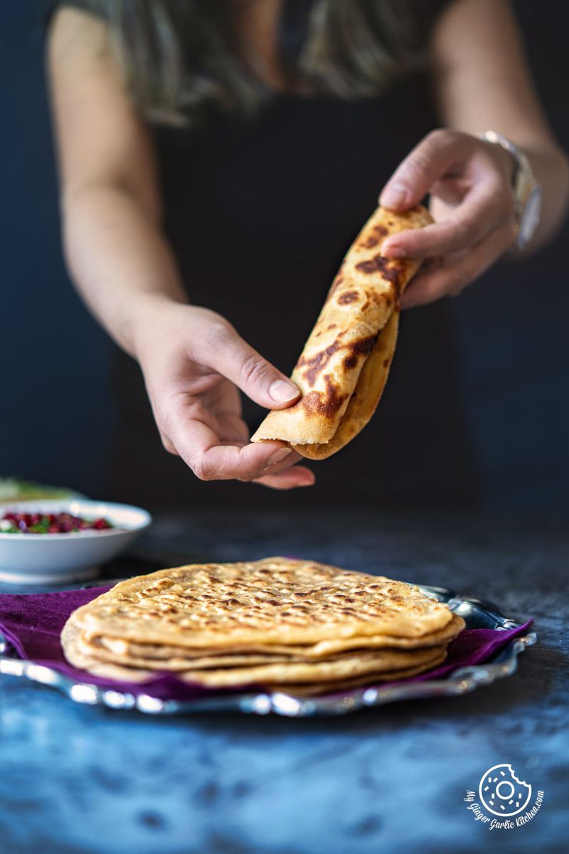 photo of a woman is holding a piece of paratha over a plate of triangle shaped parathas
