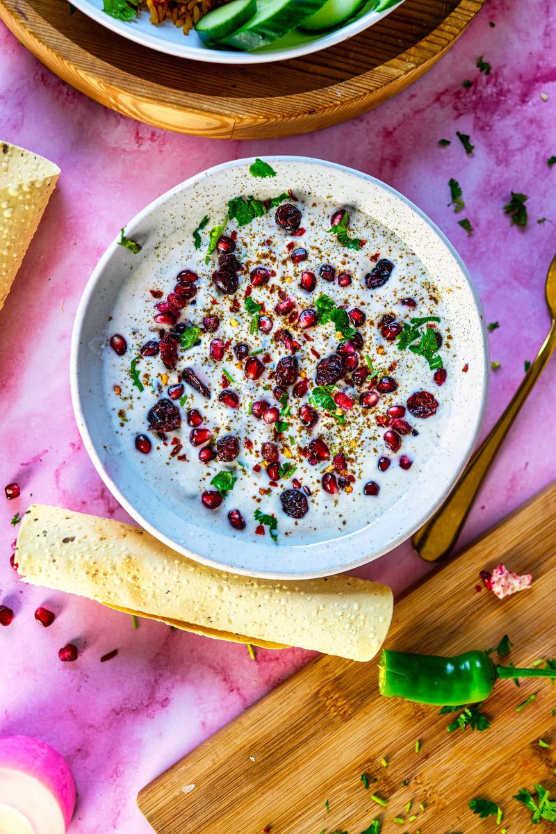 Bowl of creamy Pomegranate Raita garnished with pomegranate arils, dried cranberries, and fresh cilantro, served with a papadum and green chili on a pink surface.