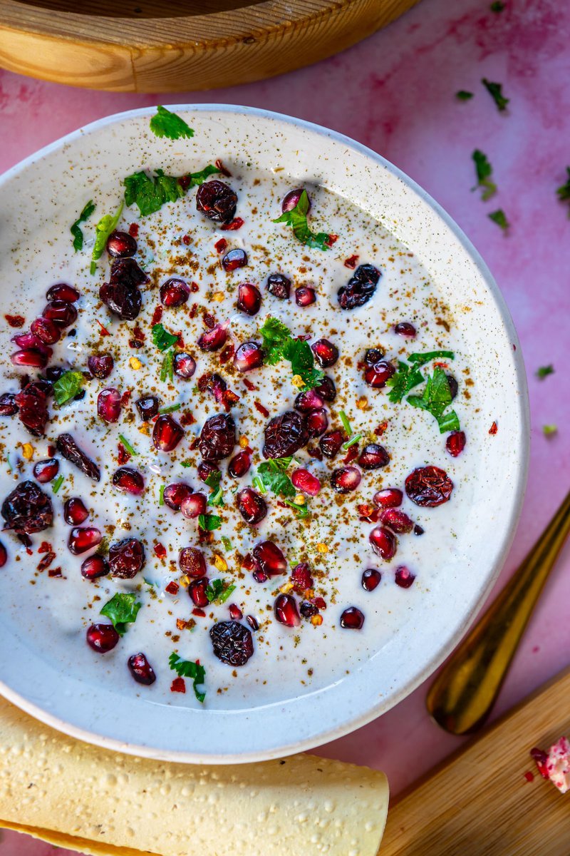 Close-up of Pomegranate Raita topped with pomegranate seeds, dried cranberries, and cilantro, showcasing a mix of creamy yogurt and vibrant red arils on a pink background.