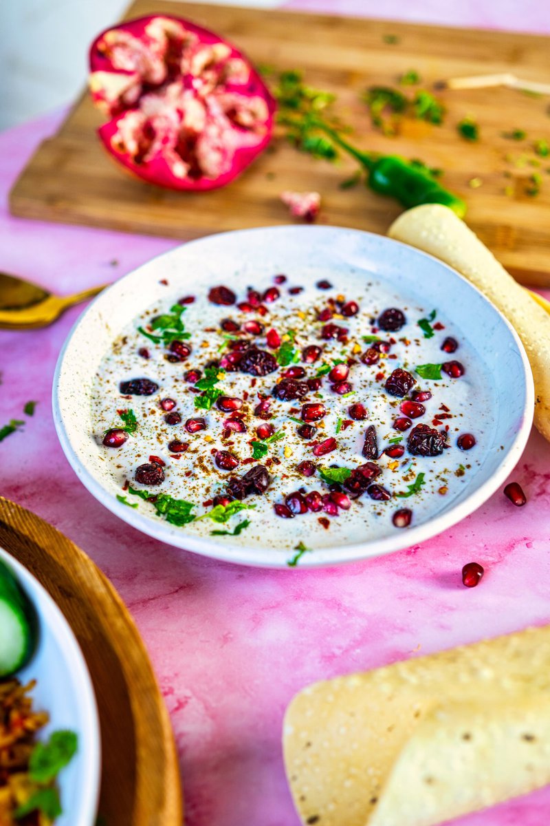 Pomegranate Raita served in a white bowl, garnished with pomegranate seeds, dried cranberries, and cilantro, with a halved pomegranate and green chili in the background.