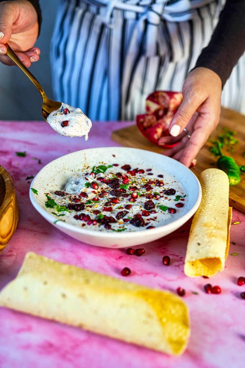 Person holding a spoonful of Pomegranate Raita above a bowl filled with the raita, garnished with pomegranate seeds, dried cranberries, and cilantro, with papadum on the side on a pink surface.