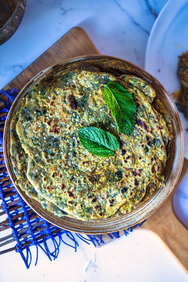 Overhead view of mint paratha (pudina paratha) in a rustic wooden bowl, garnished with fresh mint leaves. The flatbread shows its characteristic green-speckled surface with golden-brown spots from cooking. Placed on a marble countertop.