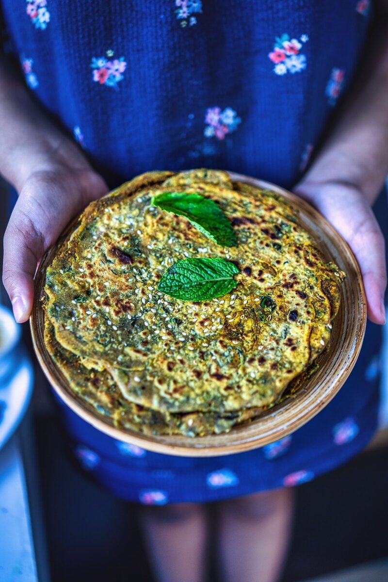 Hands holding a wooden plate with freshly made mint paratha (pudina paratha) garnished with fresh mint leaves. The green-speckled flatbread has a golden-brown surface with distinctive charred spots.