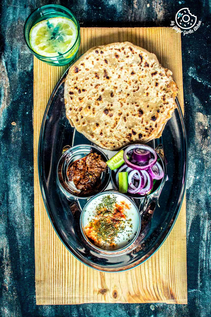 there is a plate of tandoori aloo ka paratha, pickle, raita, onions and a drink