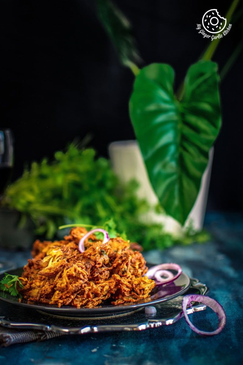pyaaz pakora served in a dark grey plate with some onion rings and some leaves in the background