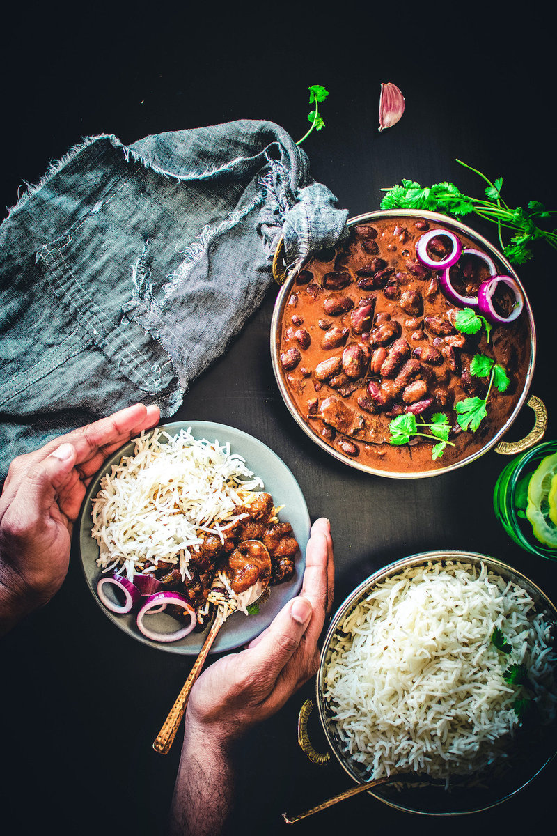A close-up of a traditional clay bowl filled with kidney bean curry topped with white rice and fresh coriander, accompanied by a rustic spoon and pieces of flatbread, all set against a dark background with vibrant green accents.