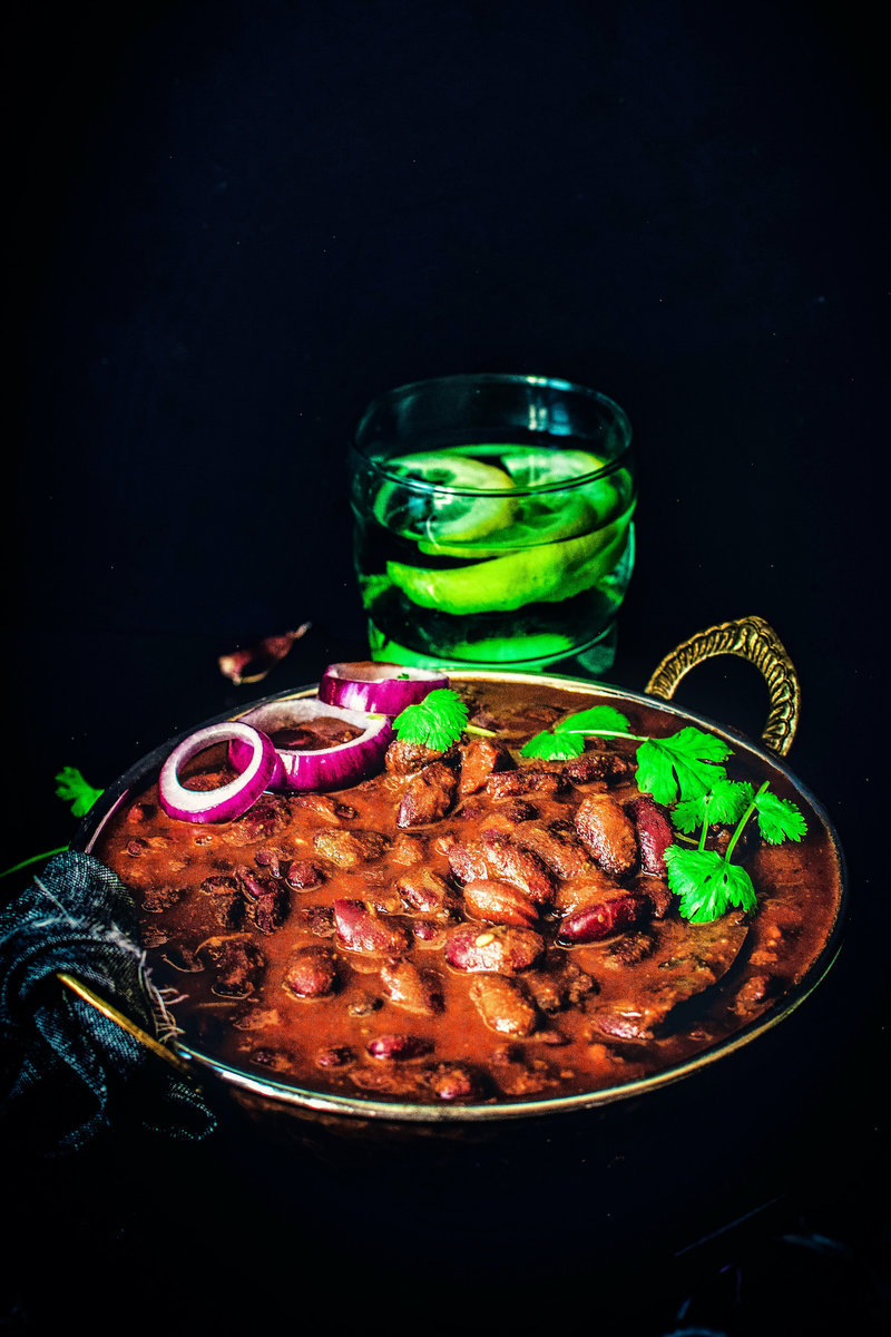 A photo of a bowl of rajma masala, a popular North Indian dish made with red kidney beans in a thick gravy. The dish is garnished with cilantro and served with a side of sliced red onions and a glass of lemon water.