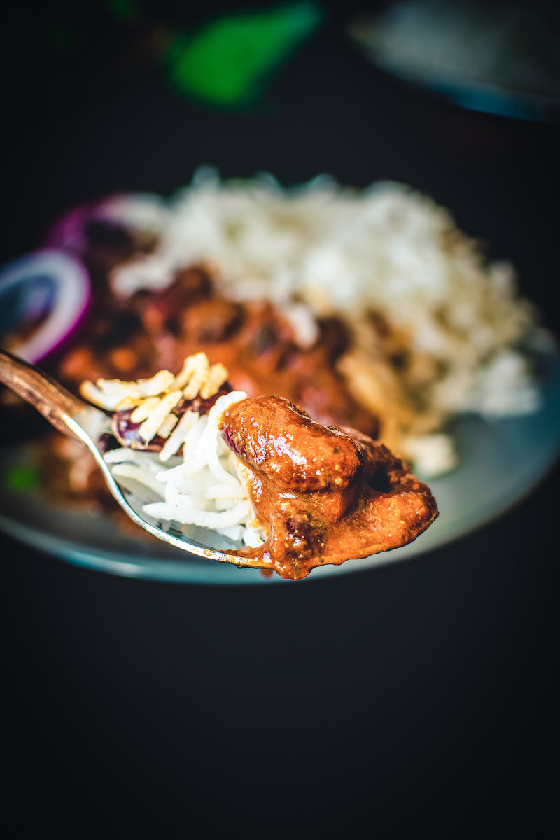 A close-up image of a spoon holding a bite of rajma masala, a popular North Indian dish made with red kidney beans in a thick gravy, served with rice.