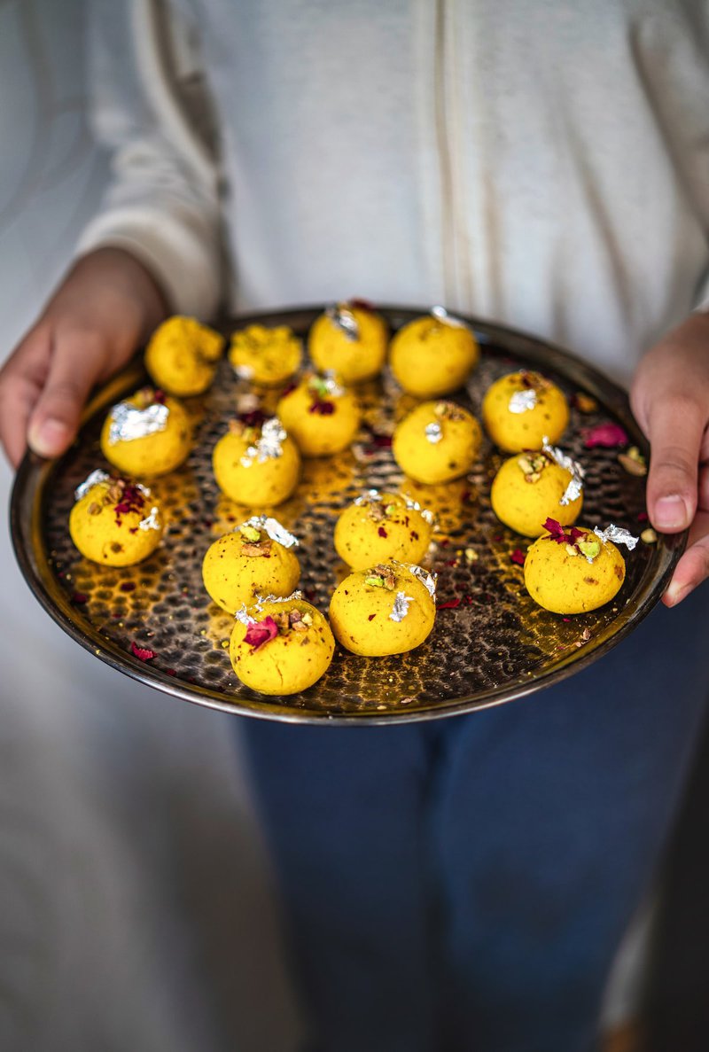 A person holding a tray of vibrant yellow Rasmalai Ladoo garnished with silver leaf and rose petals.