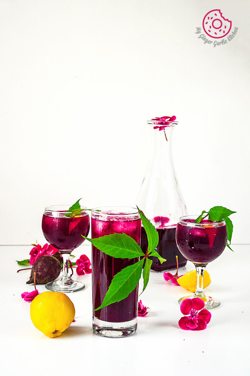 two glasses of beet lemonade with a pitcher, lemon, and a pink flowers