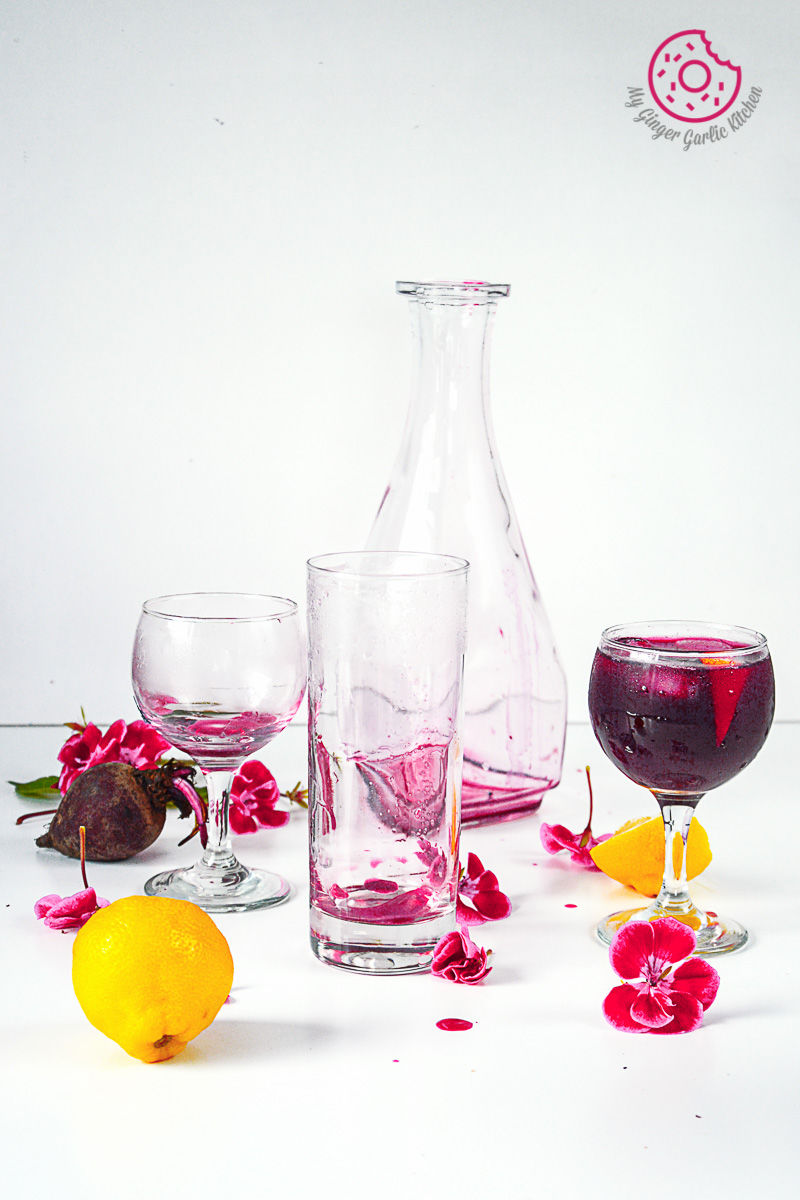 a glass of beet lemonade with a pitcher and a pink flowers on a table