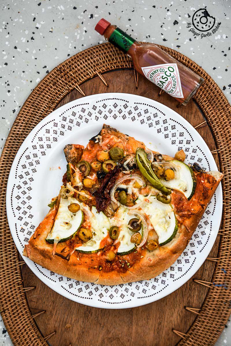 a slice of chickpea zucchini mushroom pizza with and olives on a plate with a bottle of sauce