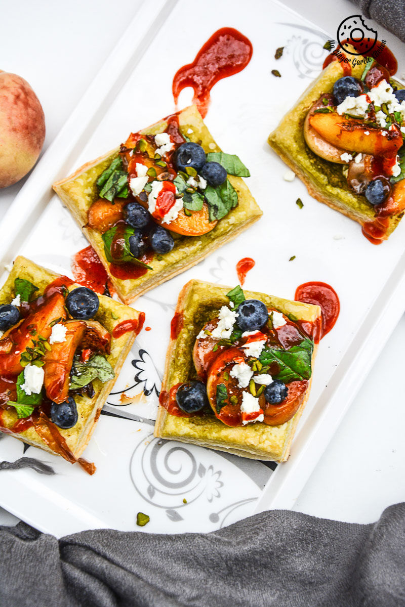 four small square peach pesto tart with blueberries and basils with fruit and cheese on a plate