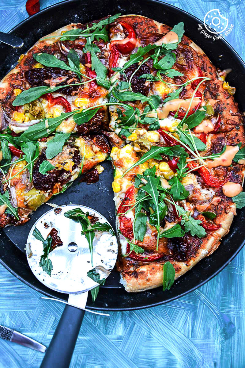 a pizza with pepper corn arugula pizza with sun dried tomatoes in a pan with a pizza cutter on a table