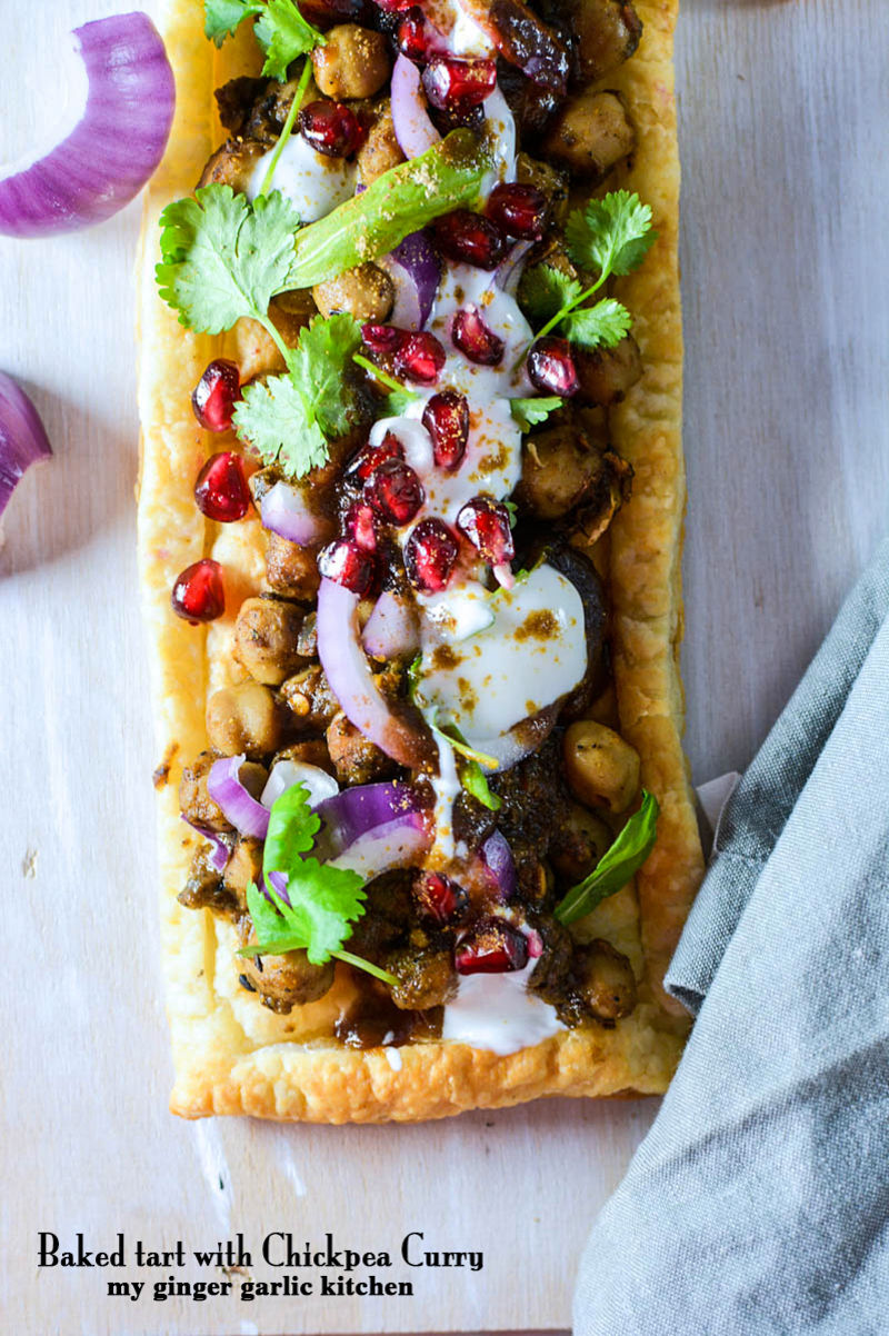 a long rectangular baked tart with chickpeas curry and tamarind chutney with chickpeas, onions, and pomegranate on it