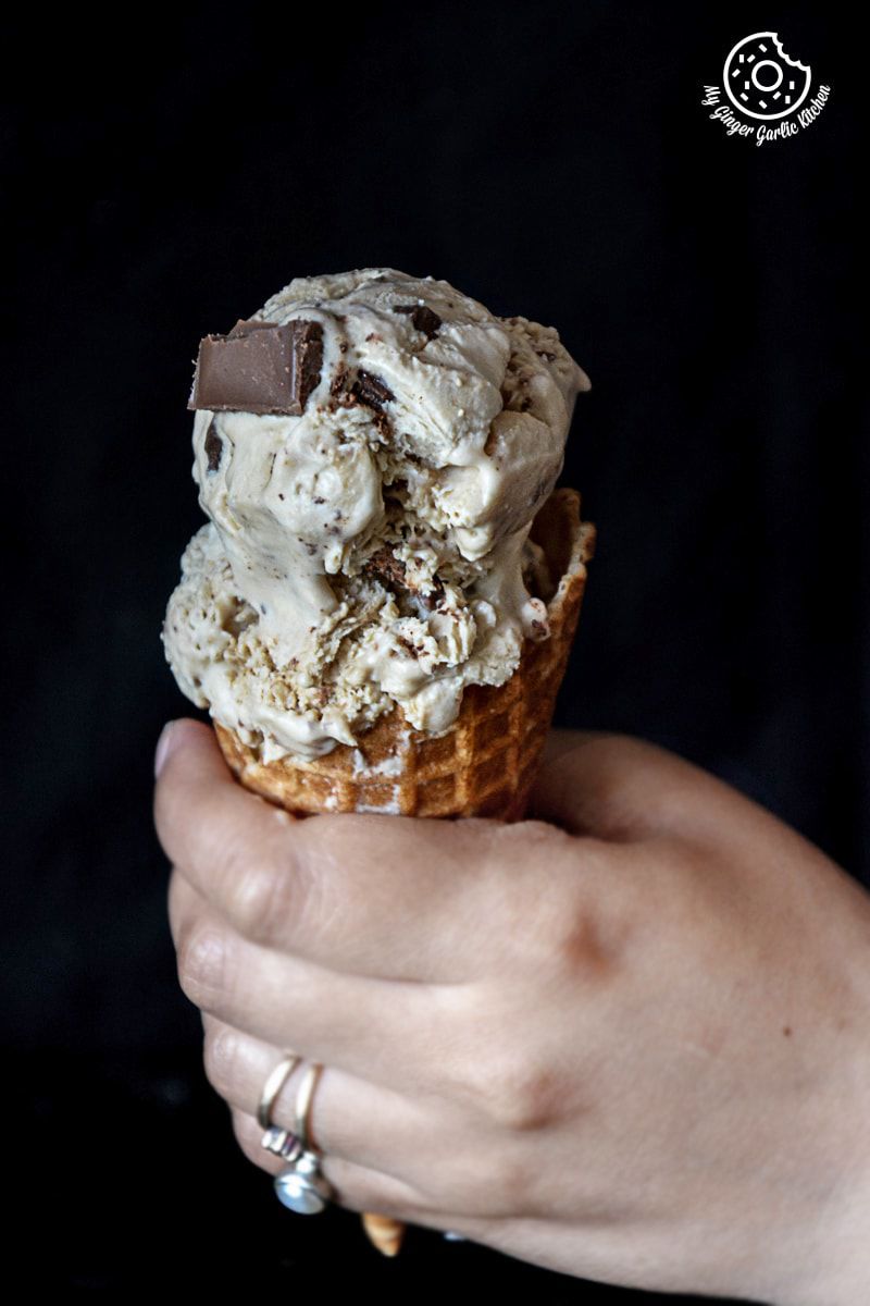 someone holding a cone of creamy coconut coffee chocolate chunk ice cream with chocolate chunks on top