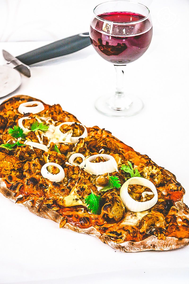 a curried cauliflower topped naan pizza with onions and mushrooms on it next to a glass of wine