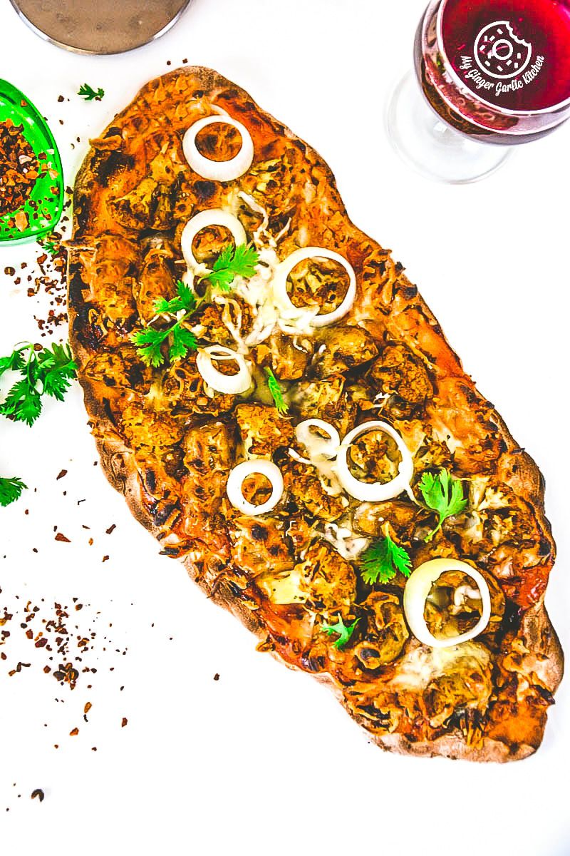 a curried cauliflower topped naan pizza with onions and herbs on it next to a glass of wine