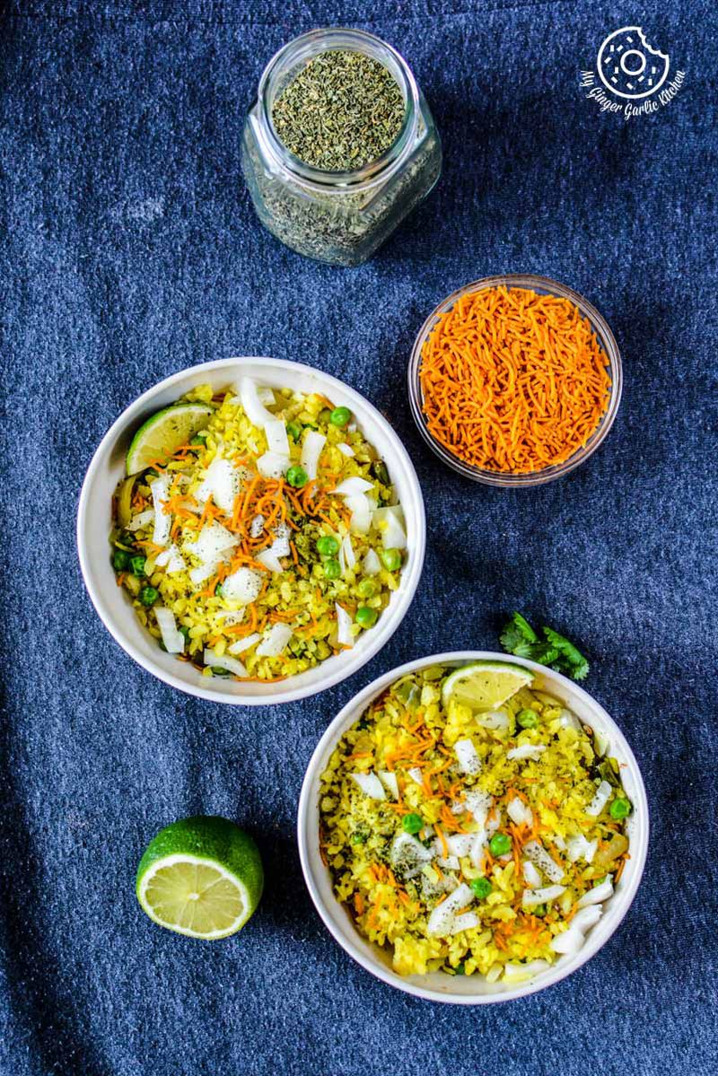 two bowls of khatta meetha indori poha with a topping of sev and cilantro, lime and sev bowl on the side