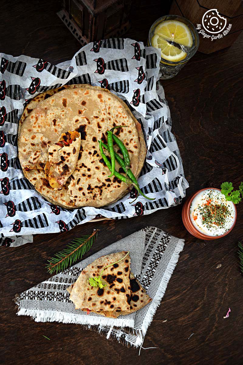 a basket of pepper onion potato paratha with green chilies on a table with a bowl of raita and a glass of lemon water
