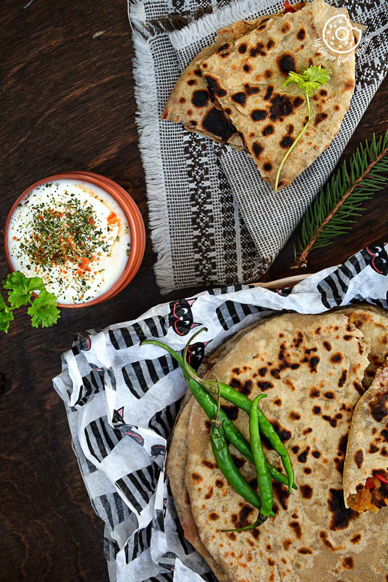 a basket of pepper onion potato paratha with green chilies with a triangle paratha on a table with a bowl of raita