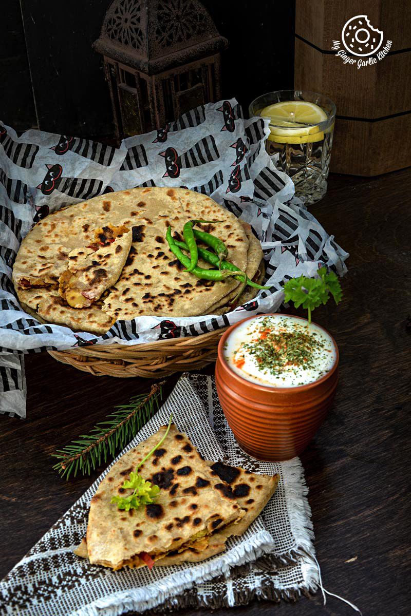 a basket of pepper onion potato paratha with green chilies with a triangle paratha on a table with a bowl of raita