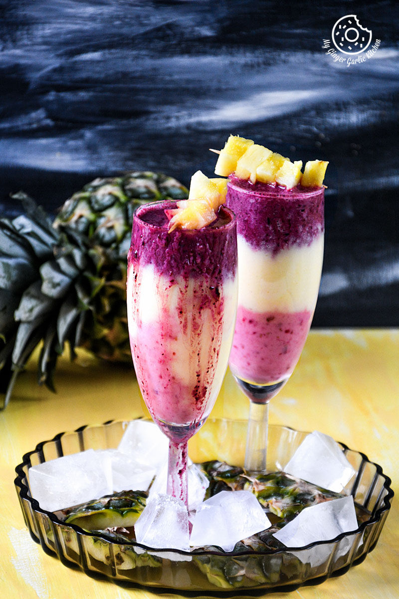 two glasses pineapple blueberry raspberry breakfast layered smoothie with ice cubes on a tray