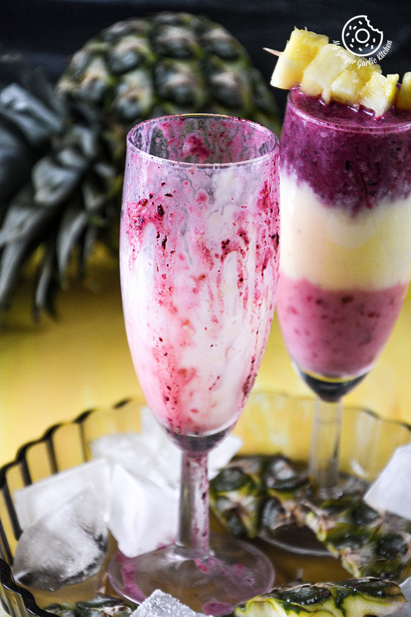 two glasses of pineapple blueberry raspberry breakfast layered smoothie and ice on a tray