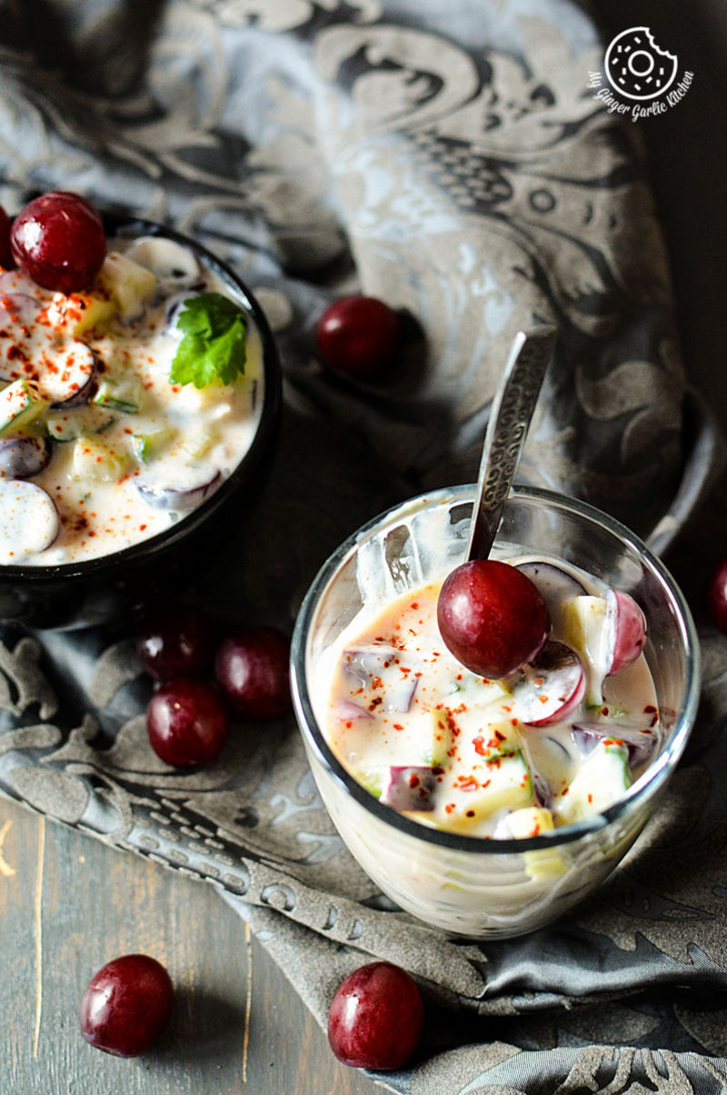 two bowls of red grapes onion cucumber raita a spoon on a table