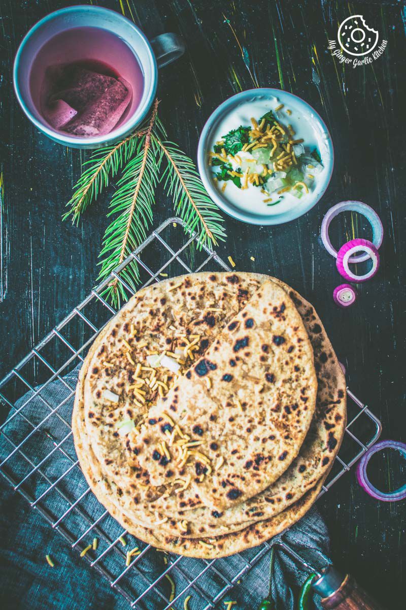 bhujia paratha stacked on a cooling rack with a bowl of raita and a a cup of tea