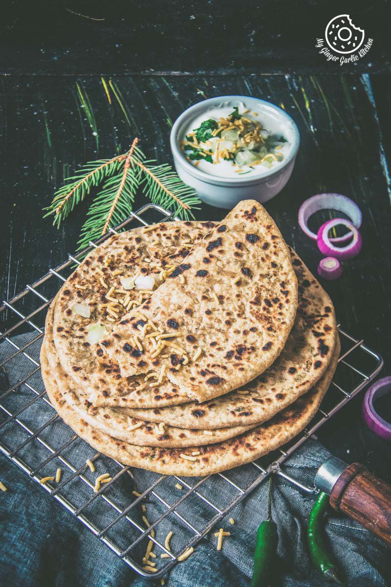 three bhujiya parathas on a cooling rack with a bowl of raita and some onions