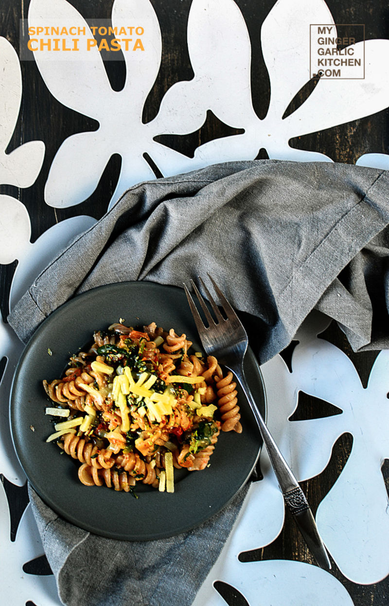 spinach tomato chili pasta dish with cheese on a grey plate with a for in it on a napkin