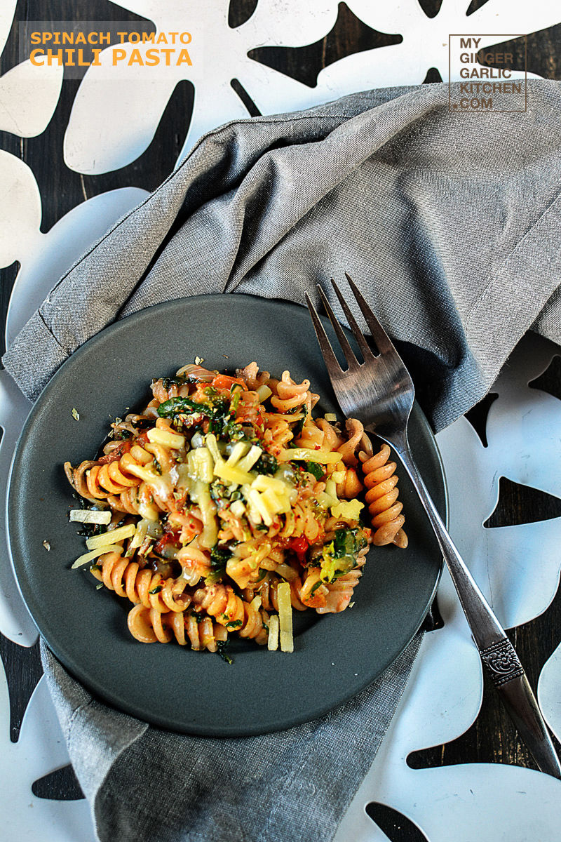 spinach tomato chili pasta dish with cheese on a grey plate with a for in it on a napkin