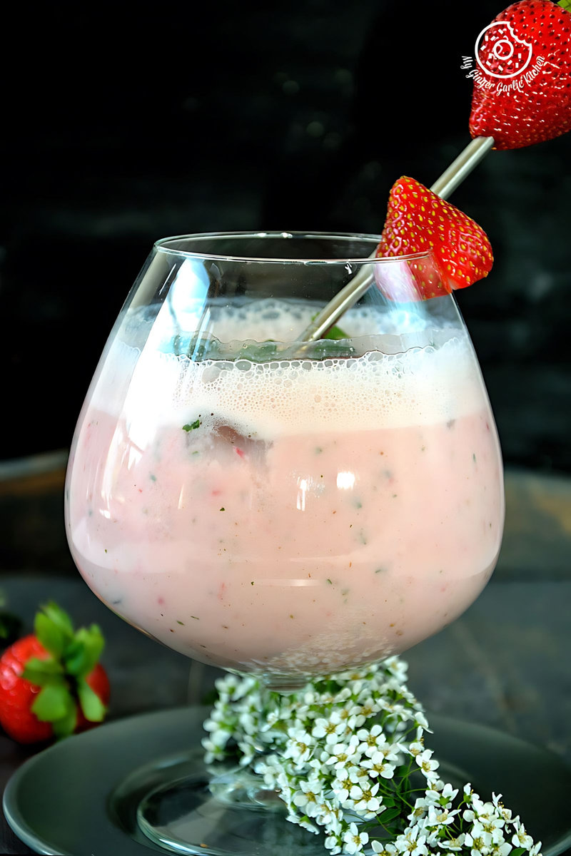 a glass of strawberry masala chaas with a straw and a strawberries on top, with some flowers