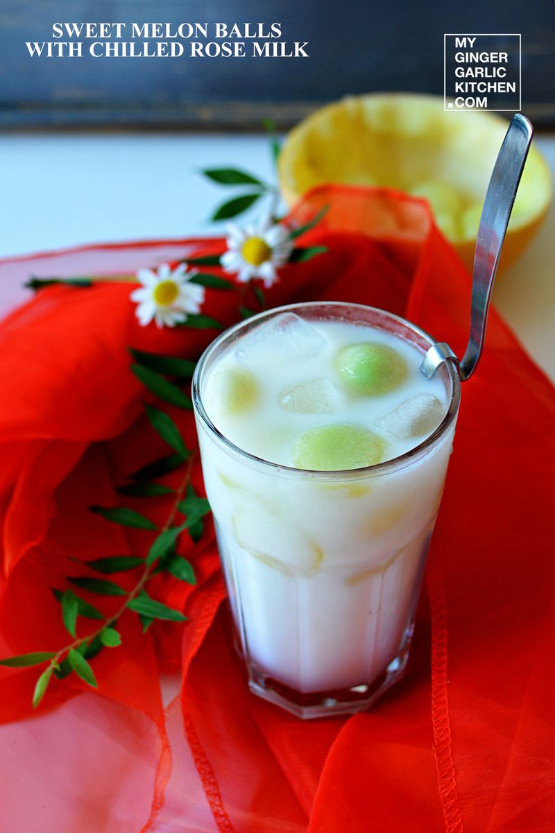 a glass of chilled rose milk with sweet melon balls some ice and some flowers with a spoon on a red cloth