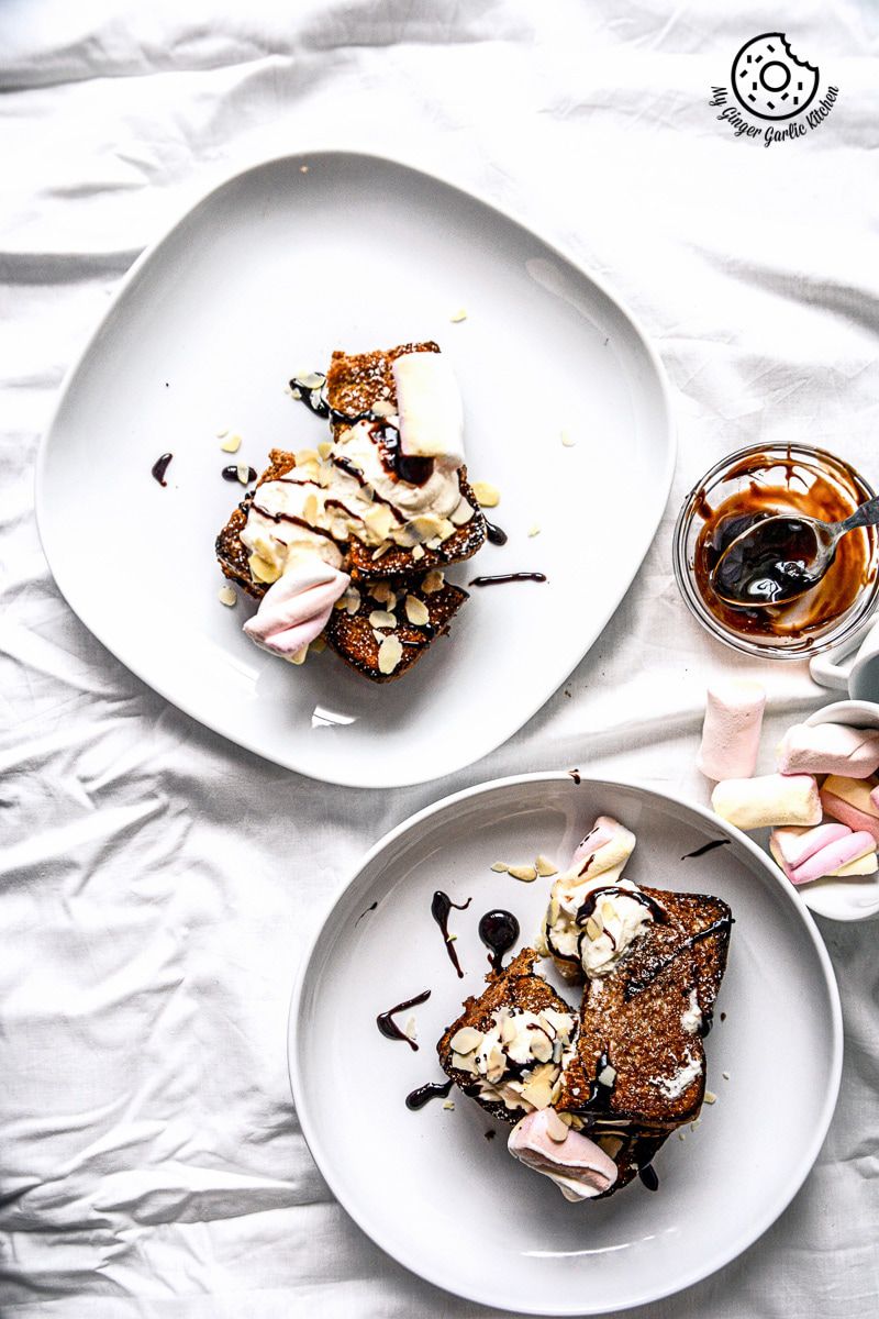 two plates of a plate with a banana nutella stuffed cinnamon maple toast whipped cream and chocolate sauce on it on a white table with a cup of coffee
