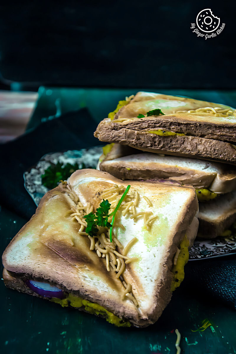 three bombay masala toasts with topped with sev and green garnish on a plate with a knife and a napkin