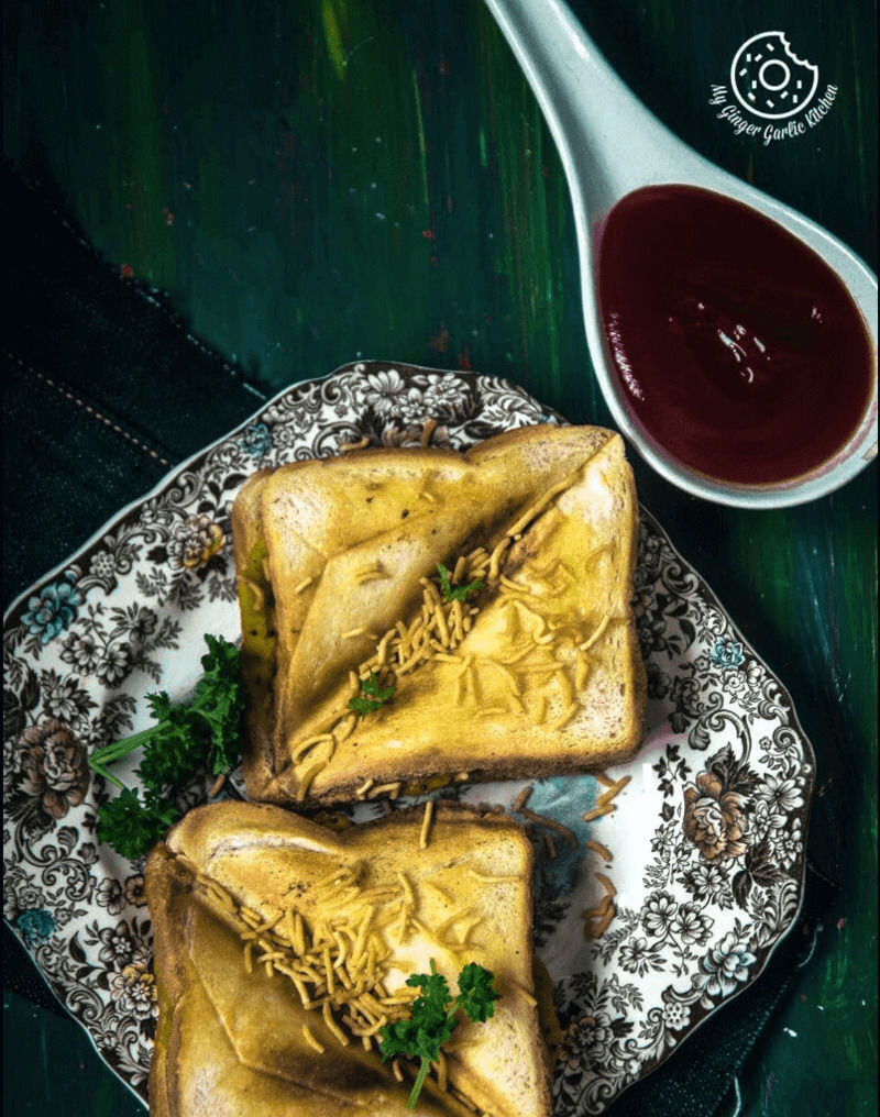 two bombay masala toasts topped with green garnish on a plate with a spoon with sauce