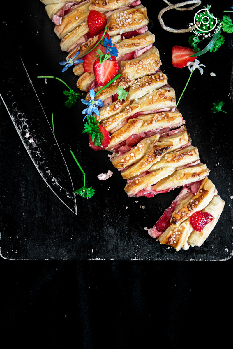 a long braided strawberry cream cheese pastry with strawberries and other toppings on a black plate