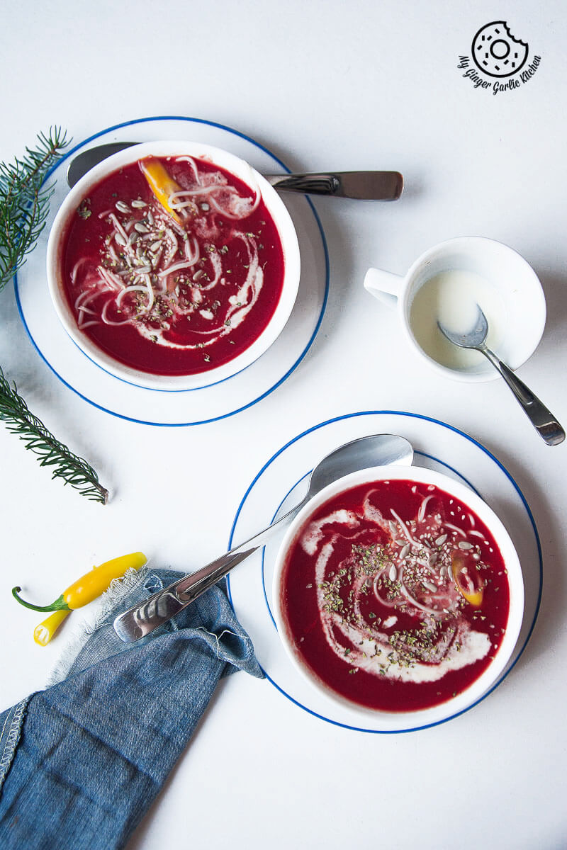 two bowls of carrot beet tomato soup on a table with a cup of cream