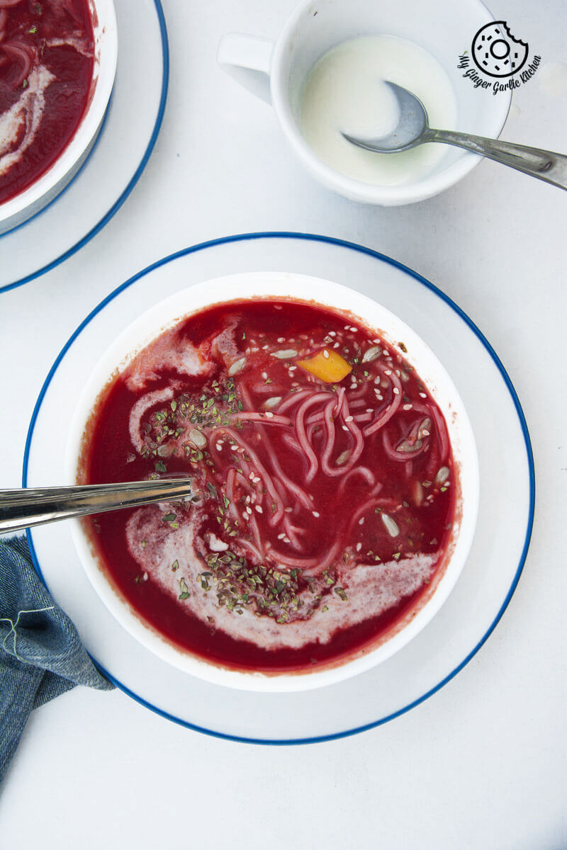 a bowl of beet tomato soup on a table with with noodles and a cup of cream