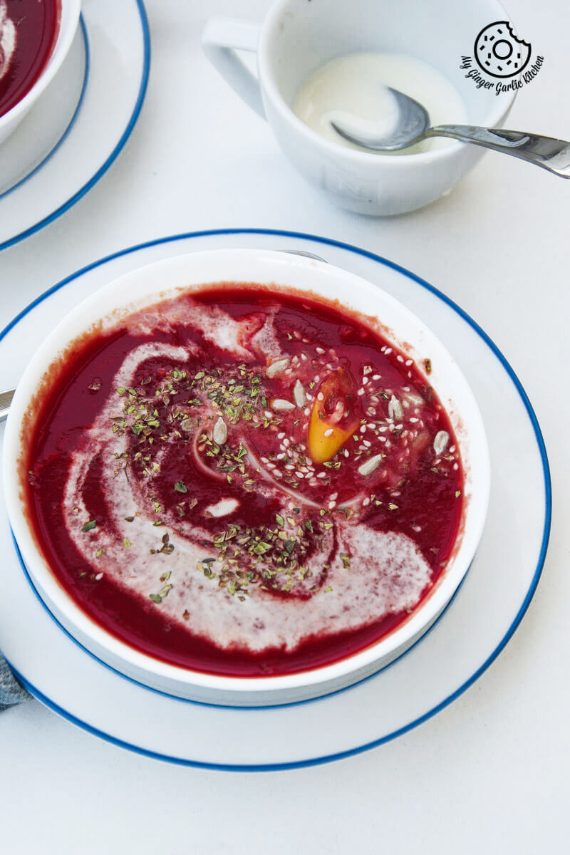 a bowl of carrot beet tomato soup topped with sesame seeds and dried herbs kept on a table with a cup of cream