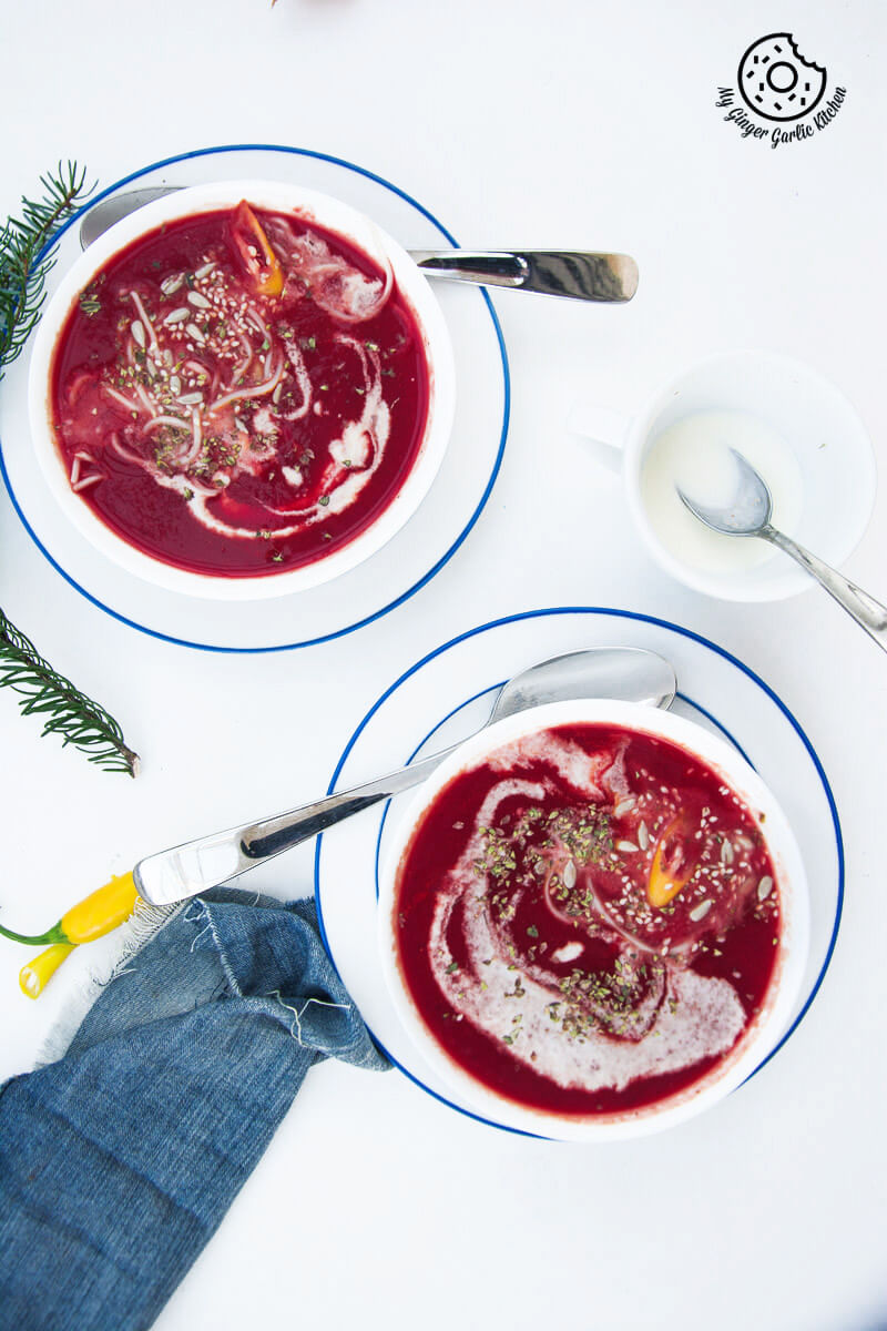 two bowls of carrot beet tomato soup topped with sesame seeds and dried herbs on a table with a cup of cream