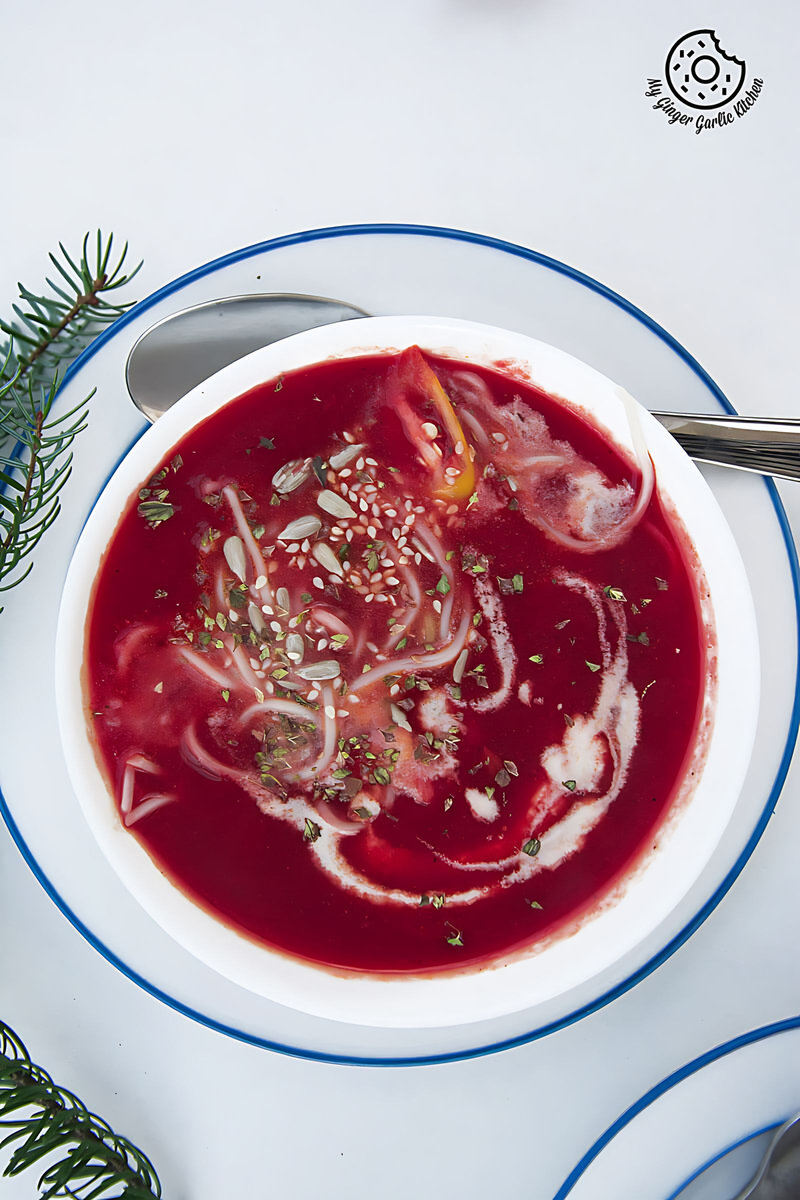 a bowl of beet tomato soup topped with noodles, esame seeds and dried herbs kept on on a plate