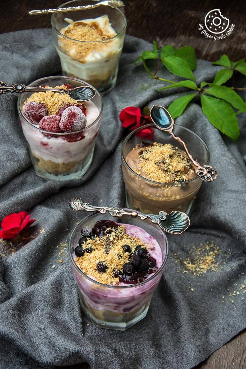 four glasses of different cheesecake shots with berries and granola on a table